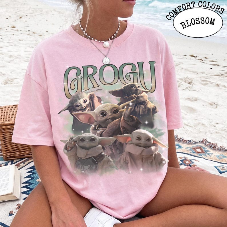 May include: A light pink short-sleeve t-shirt featuring a graphic print of four small green creatures with large ears and eyes. The text GROGU is printed in a large, stylized font above the characters. The shirt has a crew neckline and a relaxed fit.