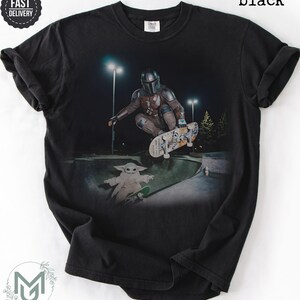 K&ouml;nnte beinhalten: Ein schwarzes T-Shirt mit einem Grafikdruck, der eine Figur in metallischer R&uuml;stung zeigt, die nachts in einem Skatepark einen Skateboard-Trick ausf&uuml;hrt, mit einem kleinen gr&uuml;nen Wesen auf einem Skateboard darunter. Der Text "black" steht oben rechts.