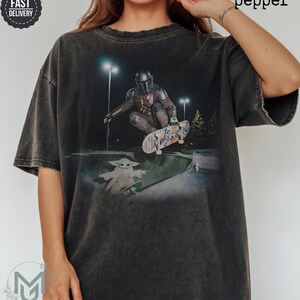 K&ouml;nnte beinhalten: Ein anthrazitfarbenes Kurzarm-T-Shirt mit einem Grafikdruck, der einen Mandalorianer zeigt, der nachts in einem Skatepark einen Skateboard-Trick ausf&uuml;hrt, w&auml;hrend eine kleine Kreatur auf einem separaten Skateboard daneben steht.