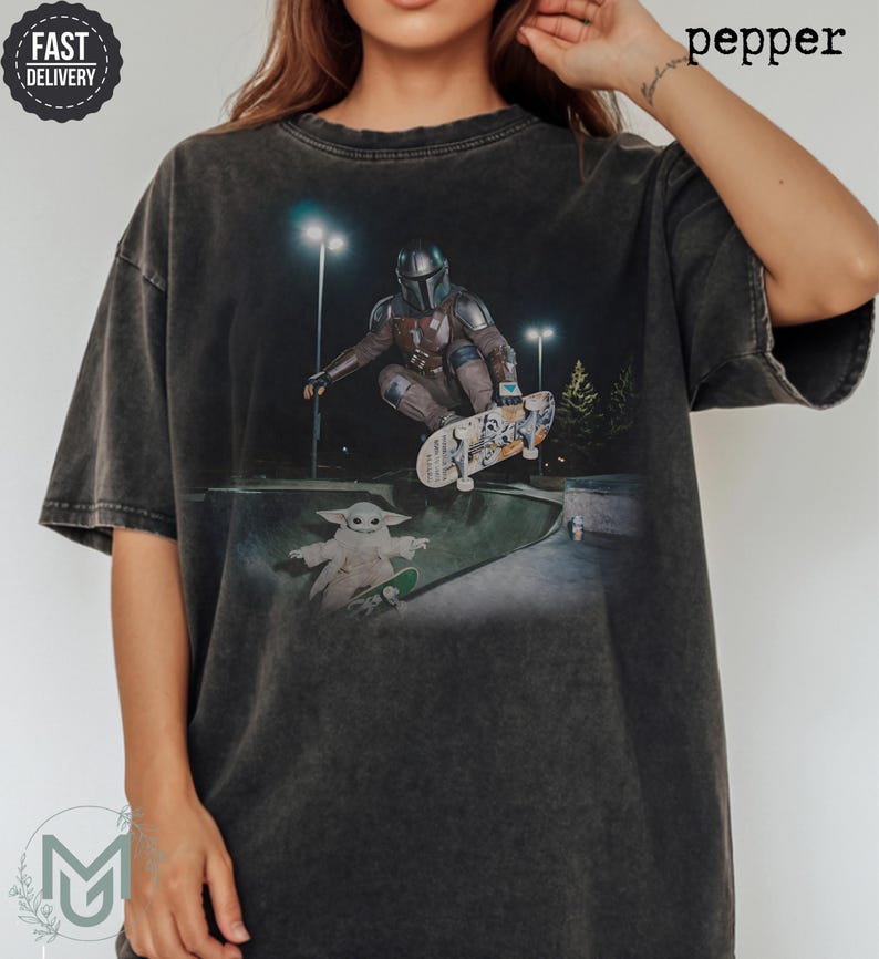 K&ouml;nnte beinhalten: Ein anthrazitfarbenes Kurzarm-T-Shirt mit einem Grafikdruck, der einen Mandalorianer zeigt, der nachts in einem Skatepark einen Skateboard-Trick ausf&uuml;hrt, w&auml;hrend eine kleine Kreatur auf einem separaten Skateboard daneben steht.