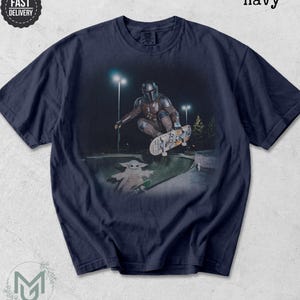 K&ouml;nnte beinhalten: Ein marineblaues T-Shirt mit einem Grafikdruck, der eine Figur mit metallischem Helm zeigt, die nachts in einer Betonsch&uuml;ssel ein Skateboard-Man&ouml;ver ausf&uuml;hrt, w&auml;hrend eine kleine gr&uuml;ne Kreatur mit gro&szlig;en Ohren auf einem Skateboard daneben steht.