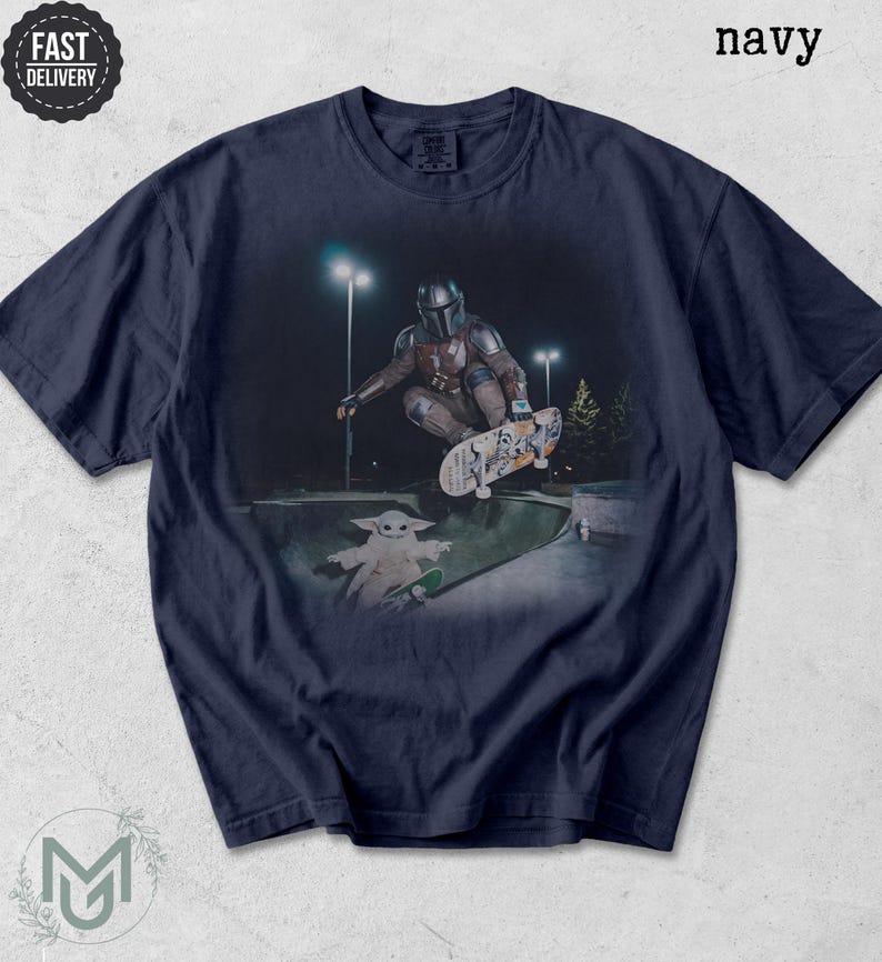 K&ouml;nnte beinhalten: Ein marineblaues T-Shirt mit einem Grafikdruck, der eine Figur mit metallischem Helm zeigt, die nachts in einer Betonsch&uuml;ssel ein Skateboard-Man&ouml;ver ausf&uuml;hrt, w&auml;hrend eine kleine gr&uuml;ne Kreatur mit gro&szlig;en Ohren auf einem Skateboard daneben steht.