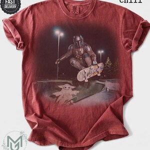 K&ouml;nnte beinhalten: Ein chilirotes Kurzarm-T-Shirt mit einem Grafikdruck, der eine Figur in Metallr&uuml;stung zeigt, die nachts in einem Park skatet, mit einer kleinen gr&uuml;nen Kreatur auf einem Skateboard darunter. Das Shirt hat einen Rundhalsausschnitt und eine Vintage-Waschung.