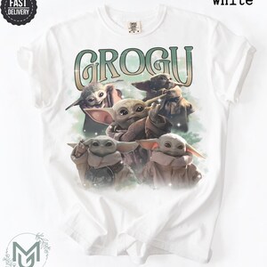 May include: A white short-sleeve t-shirt featuring a graphic print of four Grogu characters. The text GROGU is printed in a large, stylized green and white font above the characters. The design includes soft green cloud-like accents and sparkles.