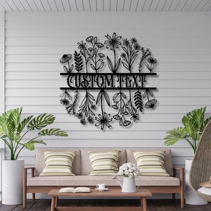 May include: Black metal floral wall art with the words "CUSTOM TEXT" in a circular design. The artwork features various flower and leaf silhouettes, perfect for indoor or outdoor decor. The piece is mounted on a light gray wall.