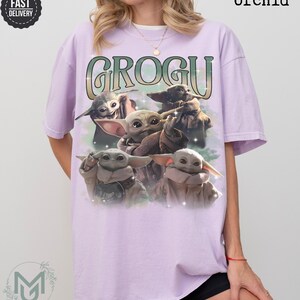 May include: An orchid-colored short-sleeve t-shirt featuring a graphic print of four Grogu characters. The text GROGU is printed in a large, stylized green and white font above the characters. The design includes subtle star-like sparkles.