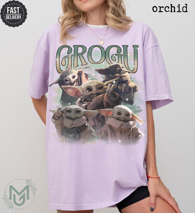 May include: An orchid-colored short-sleeve t-shirt featuring a graphic print of four Grogu characters. The text GROGU is printed in a large, stylized green and white font above the characters. The design includes subtle star-like sparkles.
