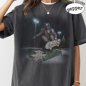 K&ouml;nnte beinhalten: Ein anthrazitfarbenes Kurzarm-T-Shirt mit einem Grafikdruck, der eine Figur in mandalorianischer R&uuml;stung zeigt, die auf einem Skateboard durch die Luft springt, &uuml;ber einer kleinen gr&uuml;nen Kreatur auf einem Skateboard. Text COMFORT COLORS pepper.