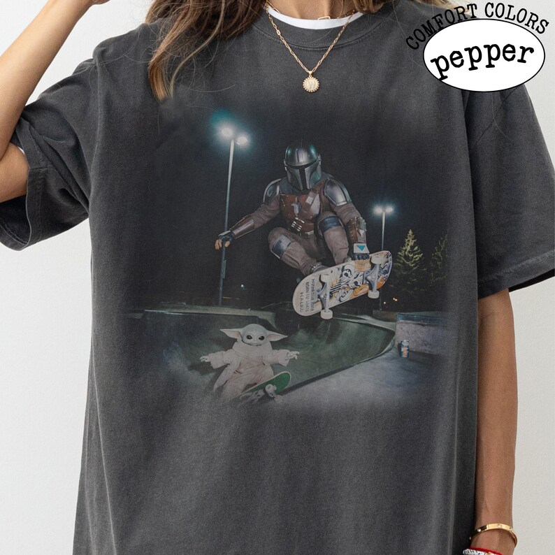 K&ouml;nnte beinhalten: Ein anthrazitfarbenes Kurzarm-T-Shirt mit einem Grafikdruck, der eine Figur in mandalorianischer R&uuml;stung zeigt, die auf einem Skateboard durch die Luft springt, &uuml;ber einer kleinen gr&uuml;nen Kreatur auf einem Skateboard. Text COMFORT COLORS pepper.