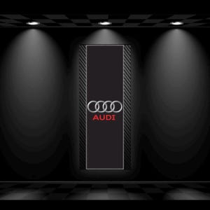 May include: A black rectangular sign featuring the Audi logo in silver and red. The sign is framed with a carbon fiber pattern and is illuminated by spotlights against a dark background. The floor has a checkered pattern.