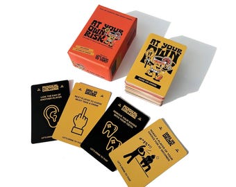May include: A set of yellow and black cards with the text "At Your Own Risk" and "Dare or Drink" on the front. The cards feature illustrations of a person drinking from a glass, a middle finger, a person removing their socks with their teeth, and a person roasting another person. The cards are part of a drinking game.