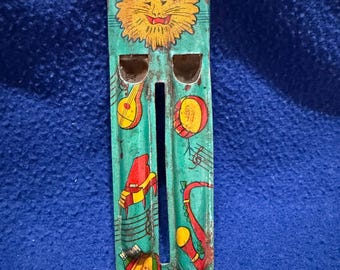 May include: A vintage tin toy whistle with a blue background and colorful musical instruments, including a saxophone, piano, and drum. The whistle features a lion's face at the top.