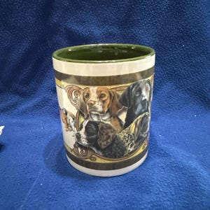 May include: A white ceramic mug with a green rim and a design of four dog heads. The dogs are a brown and white pointer, a black and white pointer, a black lab, and a brown and white springer spaniel. The design is surrounded by a gold and brown border.