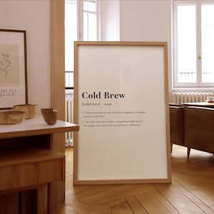 Könnte beinhalten: Ein gerahmter Druck mit den Worten "Cold Brew" in schwarzer Schrift. Die Definition von Cold Brew Coffee steht unter dem Titel. Der Rahmen ist hellbraun und der Druck wird auf einem Holzboden angezeigt.