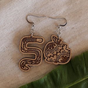 May include: A pair of wooden earrings with a detailed design. One earring is shaped like a snake with a floral pattern, and the other is shaped like an apple with a bite taken out of it. The earrings are hanging from silver hooks.