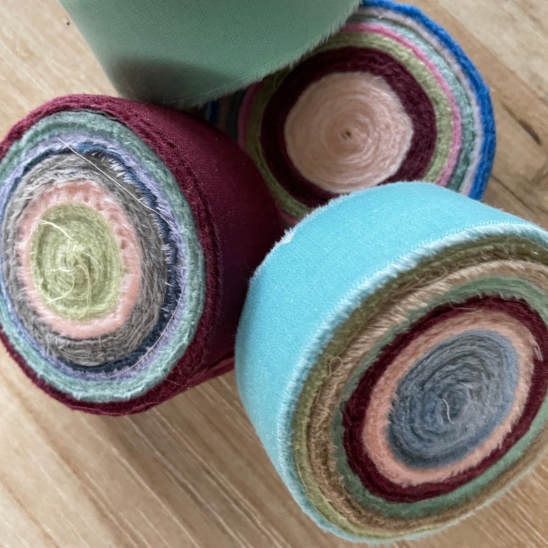 May include: Four fabric rolls, each with a different color outer layer and a multi-colored fabric roll inside. The outer layers are teal, burgundy, teal, and green. The inner rolls are all different colors, including pink, green, blue, and brown.