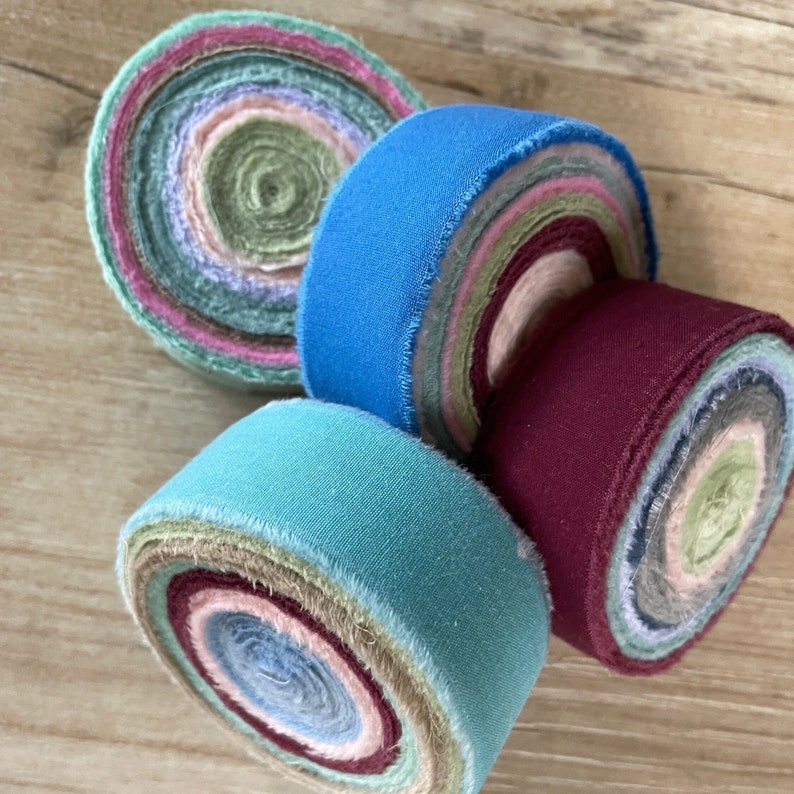 May include: Four fabric rolls, each with a different color and pattern. The rolls are made of multiple layers of fabric, creating a layered effect. The colors are teal, blue, burgundy, and a mix of green, pink, and beige.