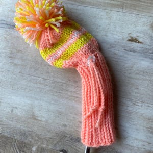 May include: A knitted golf club head cover in shades of peach, yellow, and white. The cover features a striped design and a large pom-pom on top. The cover is designed to protect the golf club head.