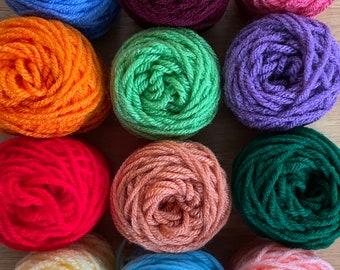 May include: Twelve balls of yarn in various colors, including red, orange, yellow, green, blue, purple, and pink. The yarn is arranged in a grid pattern on a wooden surface.