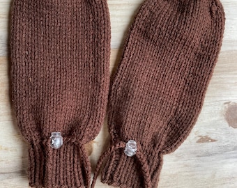 Könnte beinhalten: Ein Paar braune Strickhandschuhe mit einem Kordelzug und einem Knebelverschluss. Die Handschuhe sind aus einem weichen, warmen Garn gefertigt und perfekt, um Ihre Hände im Winter warm zu halten.