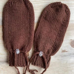 Op de afbeelding: Twee bruine gebreide wanten met een geribbelde textuur. Elke want heeft een doorzichtige plastic knoop en een trekkoord bij de pols. De wanten zijn gemaakt van een warm, zacht materiaal.