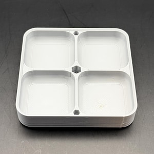 May include: A light gray, square organizer tray with four equally sized compartments. The tray has rounded corners and a raised edge. The compartments are separated by walls with a hexagonal design in the center. The tray is made of a 3D-printed material.