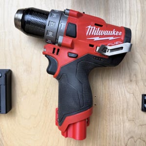 May include: A red and black Milwaukee FUEL cordless drill. The drill has a black grip, a silver chuck, and the Milwaukee logo in white. The drill is mounted on a light wood surface.