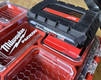 May include: A black and red Milwaukee M18 REDLITHIUM CP3.0 battery pack sits atop a red and clear plastic Milwaukee Packout storage container. The battery pack has the words "HIGH OUTPUT" printed on it.