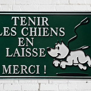 May include: A rectangular green metal sign with white lettering and a cartoon dog graphic. The sign reads "TENIR LES CHIENS EN LAISSE MERCI!" in white, with a leash attached to the dog.