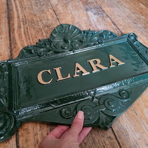 以下が含まれることがあります： 金色の文字で「CLARA」と書かれた緑色の金属製の看板。看板には装飾的で装飾的なボーダーがあり、手で持たれています。背景は木製の表面です。