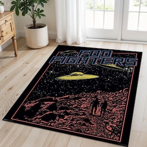 Puede incluir: Una alfombra negra y roja con un diseño espacial que presenta a la banda Foo Fighters. La alfombra tiene un fondo negro con estrellas, tres platillos voladores y dos figuras caminando sobre una superficie rocosa roja. El nombre de la banda está escrito en letras azules y rojas.