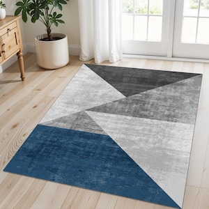 May include: Geometric area rug featuring a modern design with triangles in shades of blue, gray, and white. The rug is rectangular and placed on a light wood floor, suitable for a living room or bedroom.