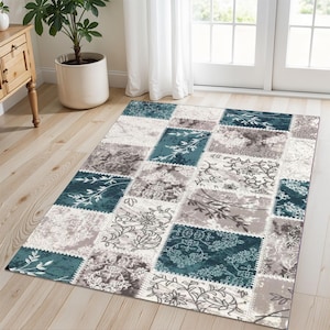 May include: A rectangular area rug with a patchwork design. The rug features squares in shades of teal, gray, and cream, with floral and leaf patterns. The rug is placed on a light-colored wooden floor, with a glimpse of a wooden cabinet and a potted plant in the background.