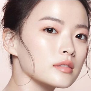 May include: Close-up of a woman's face with smooth, flawless skin. Her eyes are brown and her lips are a soft pink.