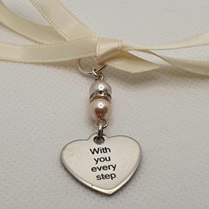 May include: A silver heart pendant with the words "With you every step" engraved on it. The pendant is adorned with two pearls and a cream-colored ribbon.