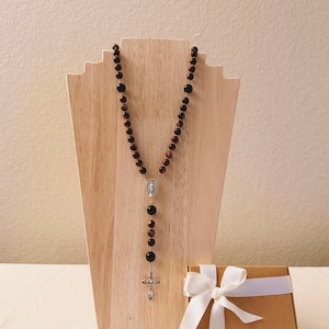 May include: A rosary with brown beads and a silver crucifix is displayed on a wooden stand. A small, brown gift box with a white ribbon is next to the rosary. The beads are a mix of dark brown and reddish-brown.