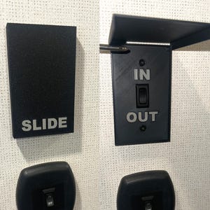 May include: Black rectangular switch plate with the word "SLIDE" in silver lettering. Another black switch plate has "IN" and "OUT" in silver lettering, with a toggle switch. The image shows a set of electrical components.
