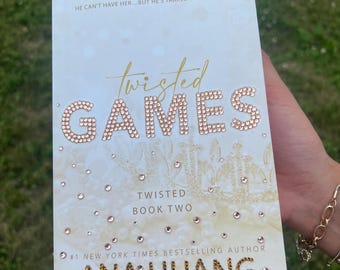 以下が含まれることがあります： アナ・フアン著「Twisted Games」の表紙。タイトルは、白い背景に大きなキラキラ光る文字で書かれています。著者の名前もキラキラ光る文字で書かれています。この本は「Twisted」シリーズの「Book Two」です。