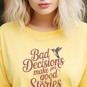 Peut inclure: T-shirt jaune avec un graphique de colibri et le texte "Bad Decisions make good Stories".