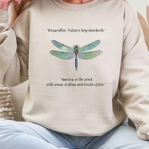 Può includere: Una felpa beige chiaro con una grafica di libellula e il testo "Dragonflies: Nature's tiny daredevils." e "dancing on the wind with wings of glass and hearts of fire."