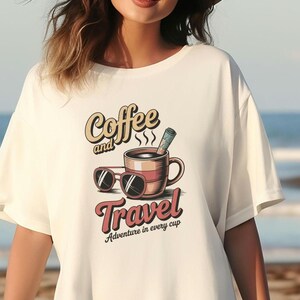 Op de afbeelding: Wit T-shirt met een grafisch ontwerp van een koffiekopje met stoom, een zonnebril en de tekst "Coffee and Travel" in een vintage lettertype. De tekst onder de afbeelding zegt "Adventure in every cup."