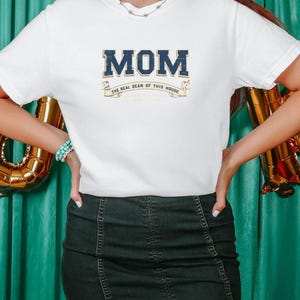 May include: White t-shirt with a blue and white graphic that says "MOM The Real Dean Of This House".