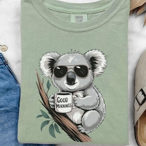 Op de afbeelding: Een lichtgroen T-shirt met een cartoonkoala die een zonnebril draagt en een mok vasthoudt met de tekst "Good Mornings".