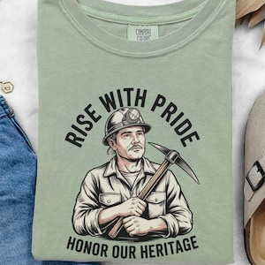 May include: A green t-shirt with a graphic of a miner holding a pickaxe. The text on the shirt reads "Rise with Pride Honor Our Heritage".