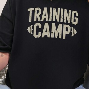 May include: Black t-shirt with a white graphic print that says "TRAINING CAMP" with a barbell on either side of the word "CAMP".