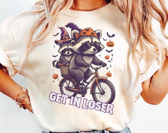 May include: Cream-colored t-shirt featuring a Halloween-themed graphic. Two raccoons are riding a bicycle, one wearing sunglasses and a hat, the other in a basket. The text "GET IN LOSER" is below the image, with bats and pumpkins.