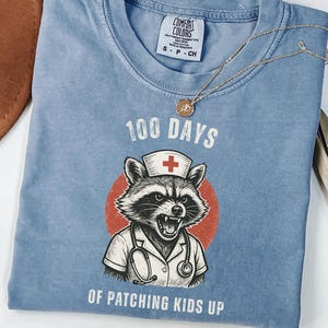 May include: A blue Comfort Colors t-shirt with a graphic of a raccoon wearing a nurse's hat and stethoscope, set against a red circle. The text reads "100 Days of Patching Kids Up." A gold necklace with a pendant is draped on the shirt.