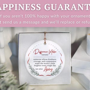 May include: A white ceramic ornament in a gift box. The ornament has the words "Difference Maker" in red script, with a definition and the name "Ashley" below. The background includes a white gift box and a sprig of greenery.