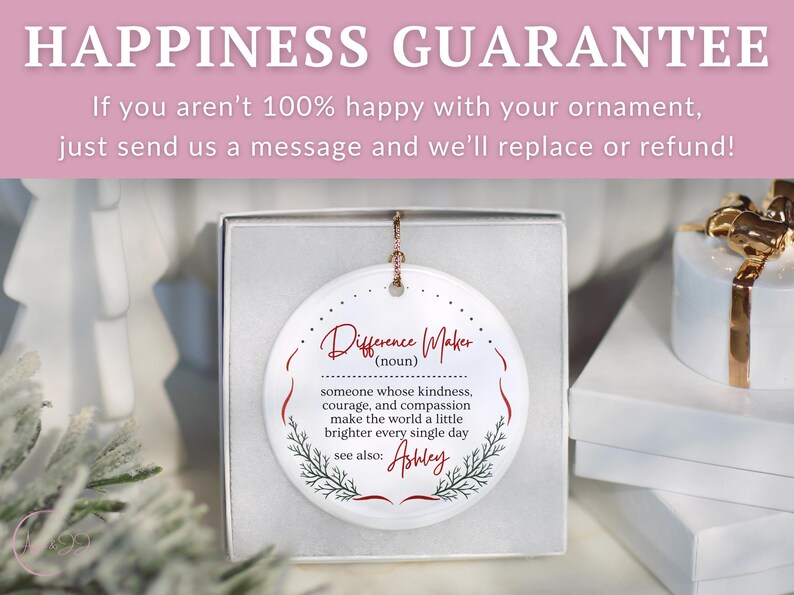 May include: A white ceramic ornament in a gift box. The ornament has the words "Difference Maker" in red script, with a definition and the name "Ashley" below. The background includes a white gift box and a sprig of greenery.