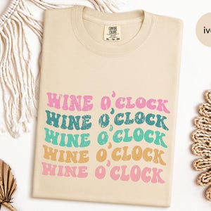 May include: A beige t-shirt with the text "Wine o'Clock" repeated five times in a retro font. The text is in shades of pink, teal, and orange.